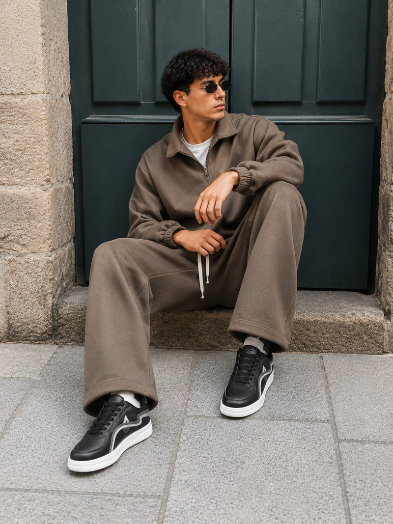 A man wearing a matching taupe brown lounge set consisting of a zip-up jacket and sweatpants, sitting on stone steps in front of a dark green door.
