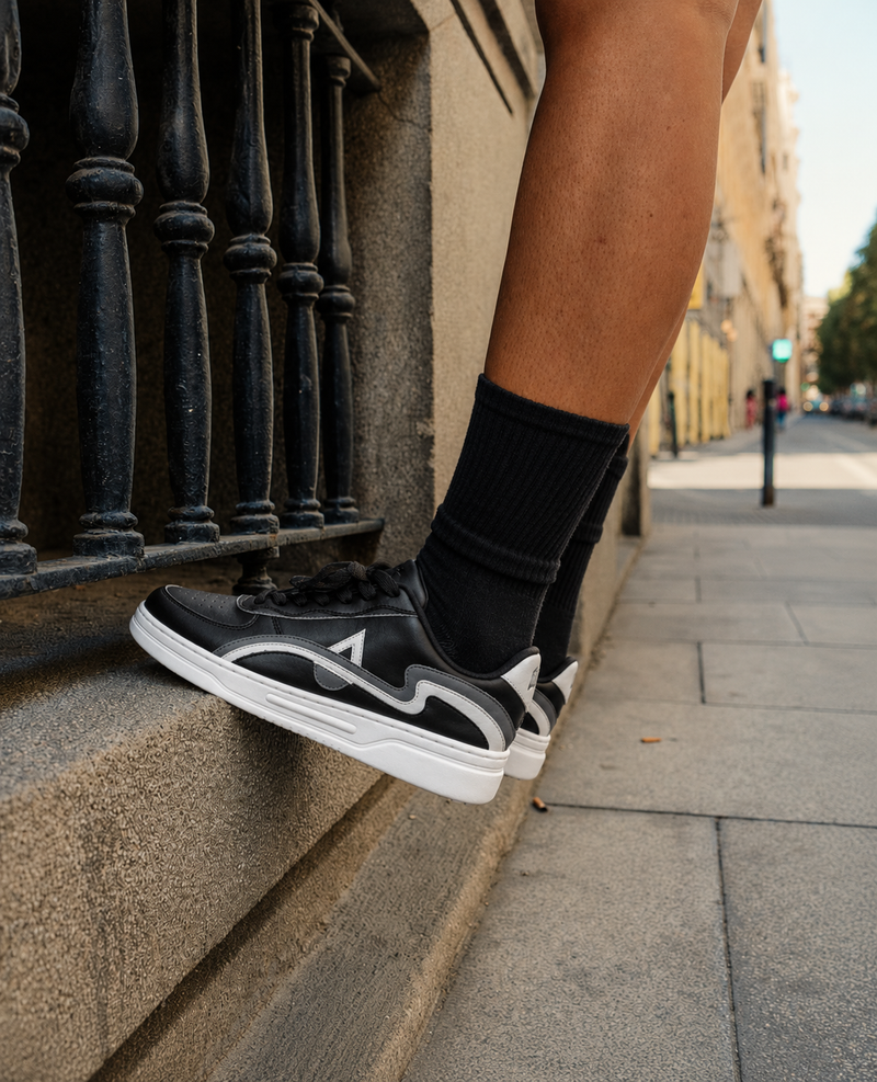 A person wearing black and white low-top sneakers with black crew socks, sitting on a stone ledge next to a black metal railing on a city sidewalk.