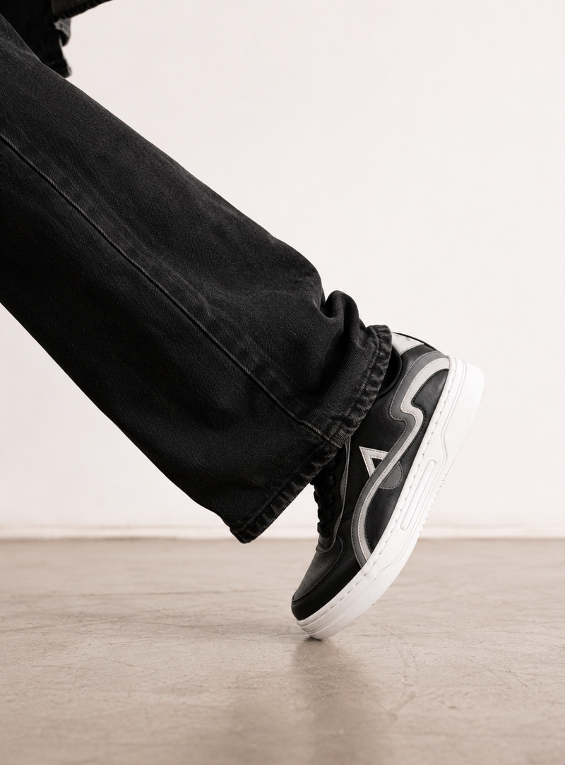 A close-up of a person's foot wearing a black and white sneaker with black jeans, captured from a low angle against a light wall and concrete floor.