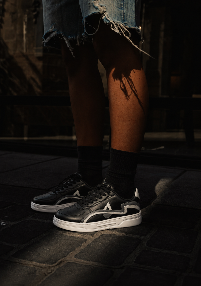 A person wearing black and white low-top sneakers with distressed denim shorts, standing on a dark cobblestone surface in dramatic low light.