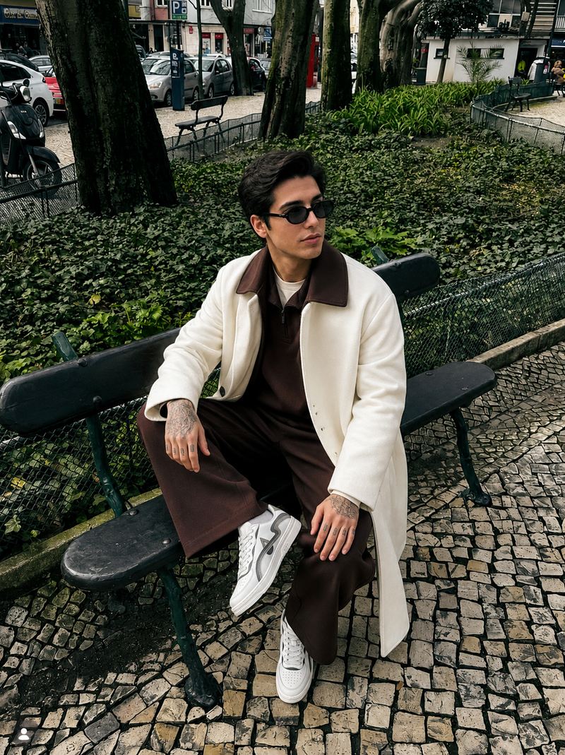 A man wearing a cream-colored overcoat with a brown collar, brown trousers, and white sneakers sits on a park bench.