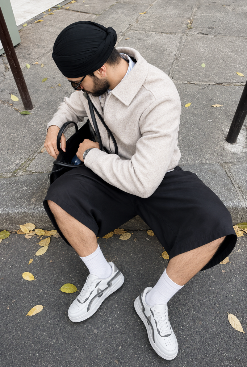 A man wearing a beige fleece jacket, black shorts, white sneakers, and a black turban sits on a stone curb outdoors, looking into a black bag.