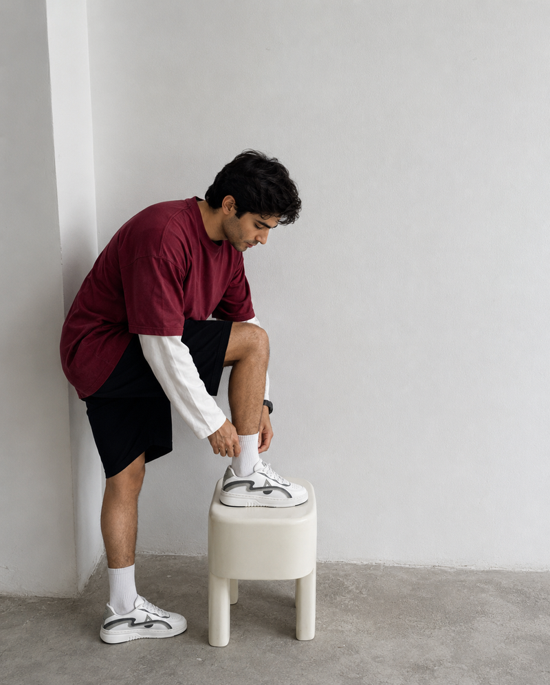 A man in a maroon t-shirt, white long-sleeve shirt, black shorts, white socks, and white sneakers with gray accents is sitting on a small white stool, tying his shoe in a minimalist room with a white wall and concrete floor.